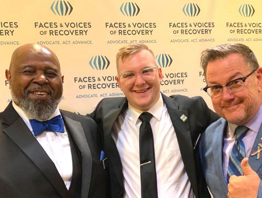 Three people in suits against Faces and Voices backdrop