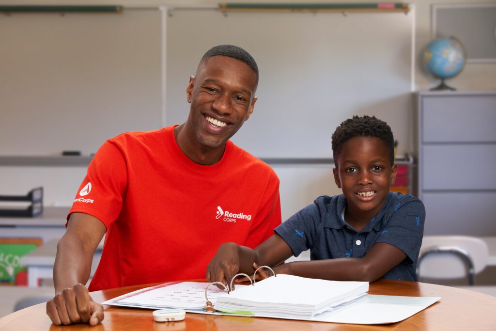 Seated side-by-side, an adult in a red shirt and a child in a blue shirt smile at the camera. They lean on a table with an open book.
