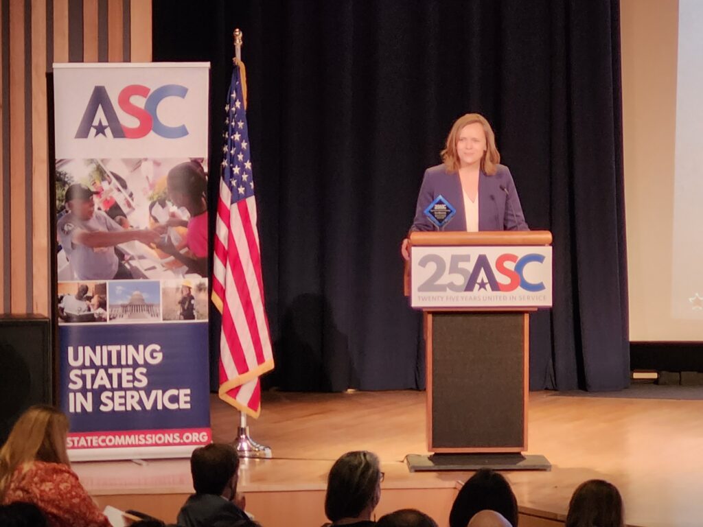 Woman stands behind podium next to a banner and American flag. Award is on top of podium.